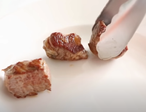 Three beef cubes cooking on a plate, being handled with tongs