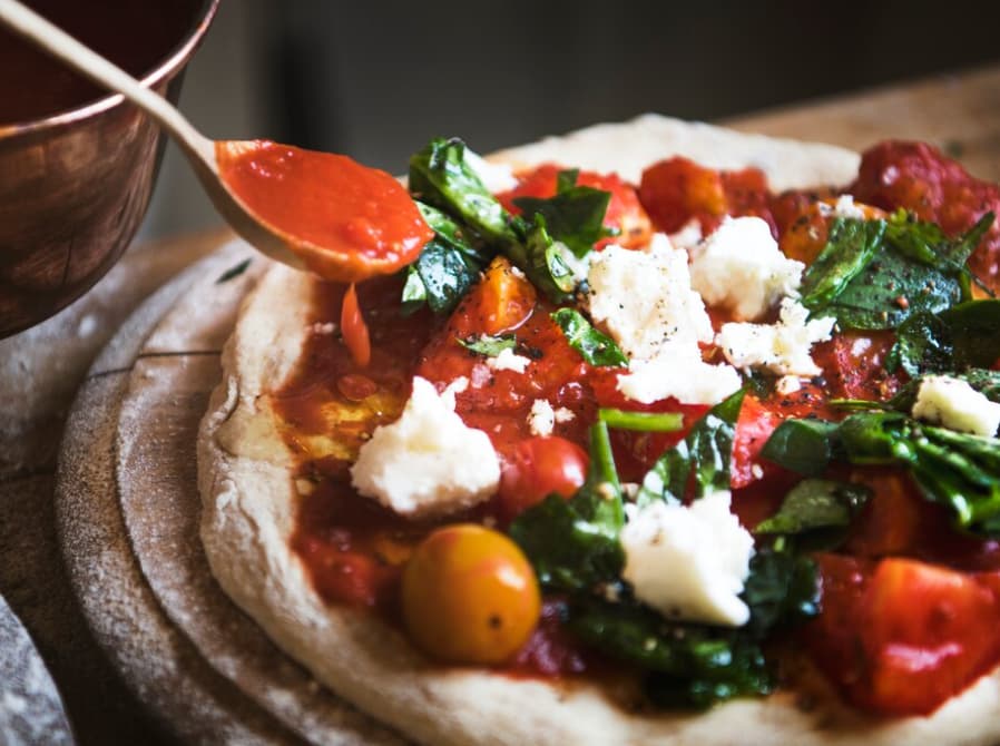 Fresh tomato sauce being spooned onto a pizza with spinach and cheese