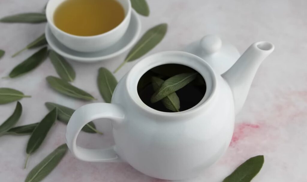 A white teapot with fresh sage leaves inside and a cup of tea on a marbled background