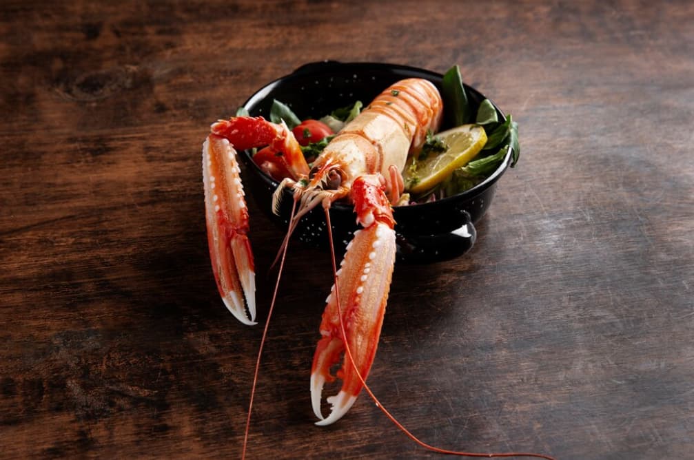 a lobster with lemon, cherry, and salad inside a black bowl on the wooden table